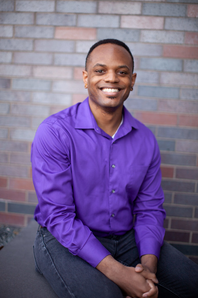 Lateef smiling while sitting on a ledge against a brick wall