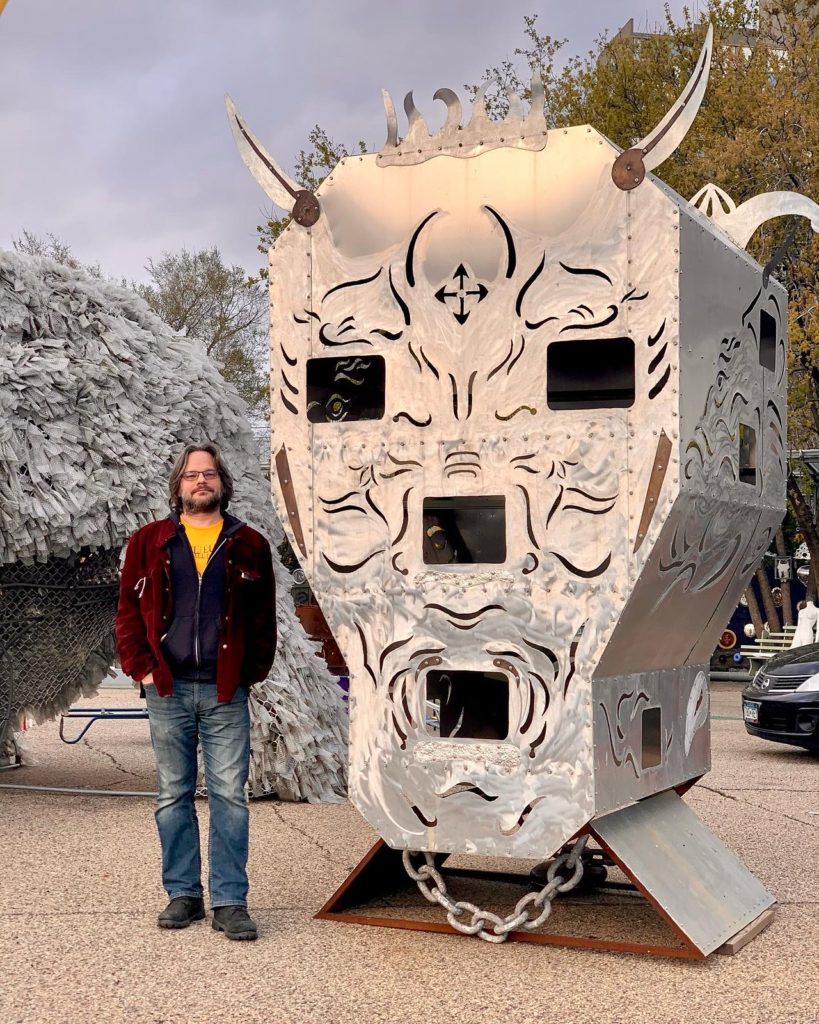 My friend James standing in front of a large metal sculpture of a head with horns