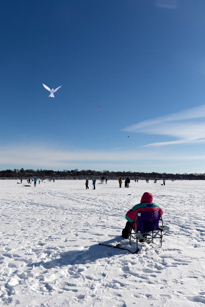 A person sitting in a chair on a frozen lake controlling a kite of a bit white dove