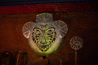 Large metal sculpture of a face hanging on a brick wall with disco lights shining on it at night