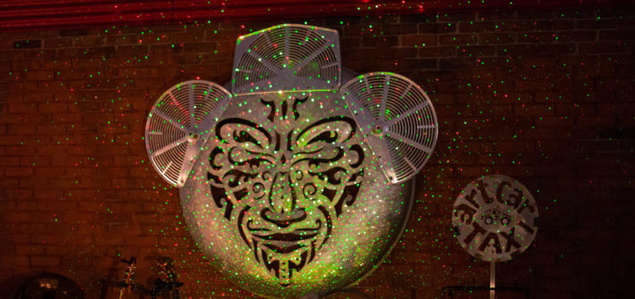 Large metal sculpture of a face hanging on a brick wall with disco lights shining on it at night