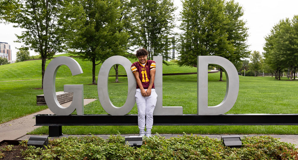 A young man in a football uniform standing with giant metal letters spelling the word GOLD