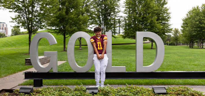 A young man in a football uniform standing with giant metal letters spelling the word GOLD